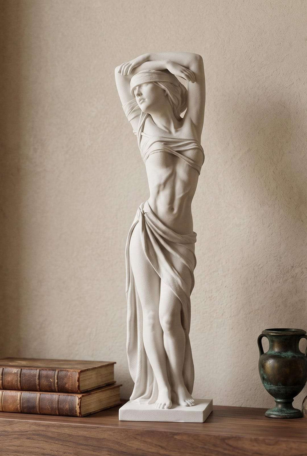 Statua bianca effetto gesso di figura femminile bendata con braccia alzate su mensola in legno accanto a libri antichi